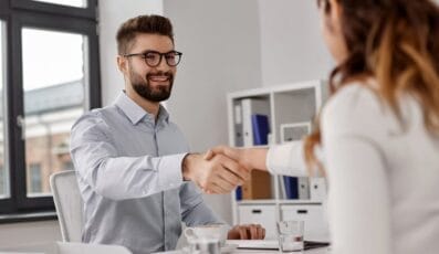 En mand og en kvinde sidder over for hinanden ved et skrivebord på et kontor og smiler og giver hånd, og foreslår en vellykket jobsøgning i Danmark eller et professionelt møde.