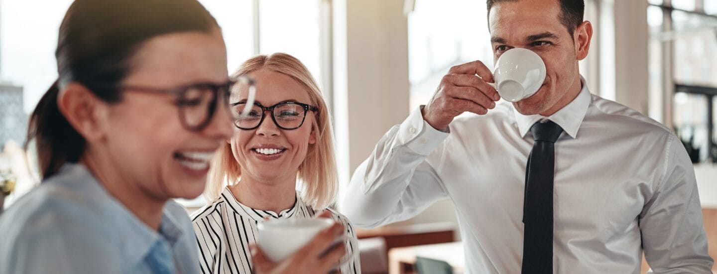 Tre professionelle i erhvervspåklædning smiler og griner over kaffen på et lyst, moderne kontor, diskuterer jobsøgning i danmark efter udalandsophold og deler tips til at komme ind på det danske arbejdsmarked igen efter en tid i udlandet.