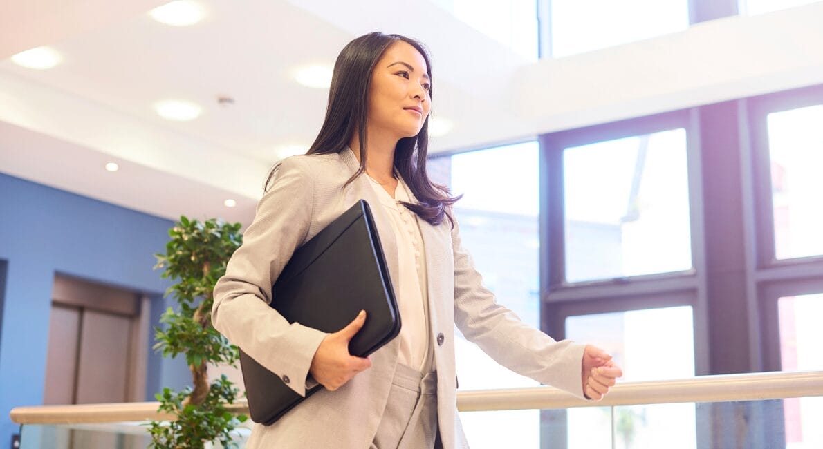 En kvinde i et lysegråt jakkesæt går selvsikkert indendørs med en sort bærbar computer under armen – og fanger ånden i jobsøgning i danmark for udlandsdanskere – med store vinduer og potteplanter i baggrunden.
