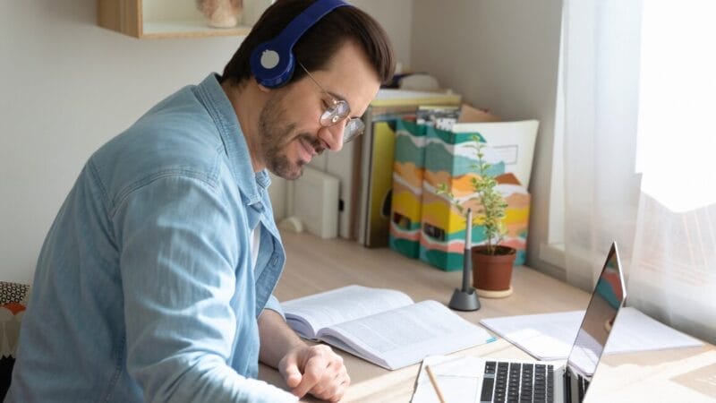 A man wearing headphones and glasses sits at a desk, smiling while writing in a notebook. An open laptop, books, and a small potted plant are on the desk near a sunlit window.