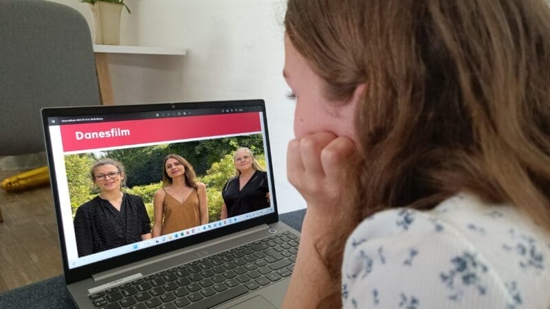 En person med langt hår kigger på en bærbar computerskærm, der viser en hjemmeside kaldet Danesfilm, med tre kvinder, der står udendørs og smiler.