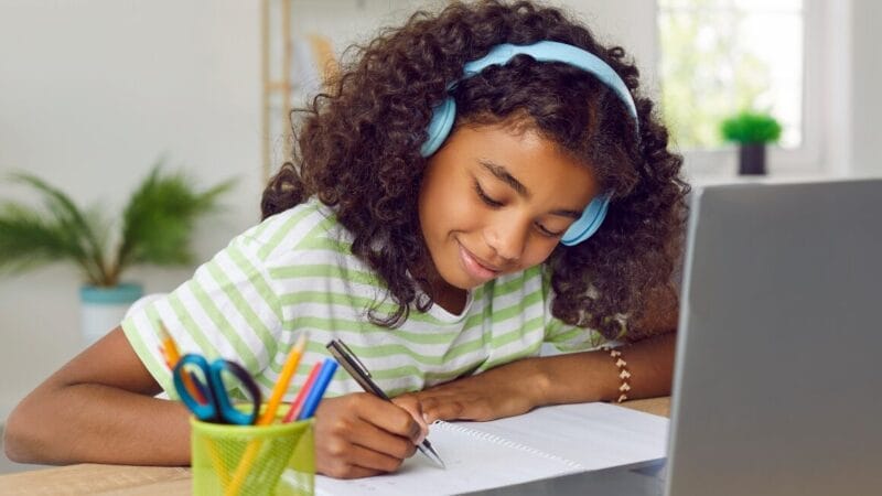 A girl with curly hair wearing blue headphones smiles while writing in a notebook, practicing Danish for beginners. She sits at a desk with a laptop, colorful pens, and a green plant in the background.