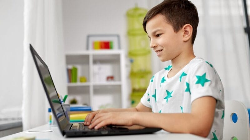 En ung dreng iført en hvid skjorte med grønne stjerner smiler, mens han bruger en bærbar computer ved et skrivebord i et lyst rum med hylder og farverige bøger i baggrunden.