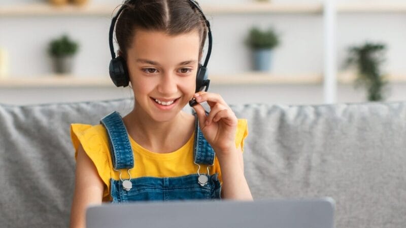 En ung pige iført en gul skjorte og denimoveralls sidder smilende i en sofa og bruger et headset, mens hun kigger på en bærbar computerskærm i hyggelige, hjemlige omgivelser.