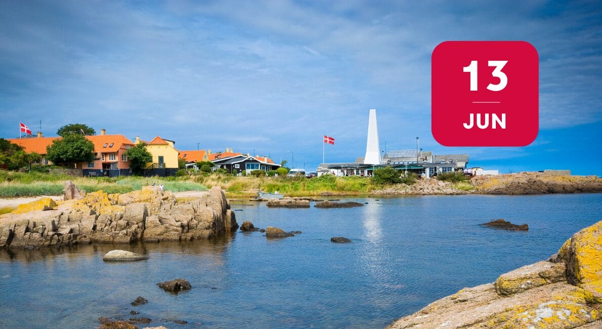 Kystlandsby med farverige huse, danske flag, klippefyldt kystlinje og et højt hvidt monument under en blå himmel. En rød firkant viser datoen 13. JUNI – hvilket markerer starten på Folkemødet 2025.