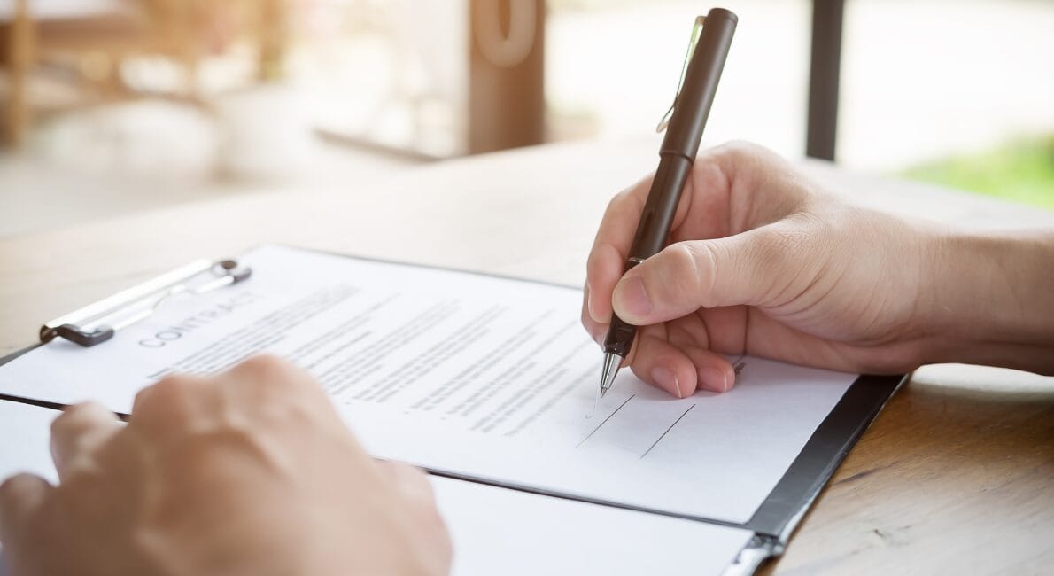 En person med en pen underskriver et Høringssvar-dokument på et udklipsholder ved et bord, hvor naturligt lys falder ind gennem et vindue i nærheden.