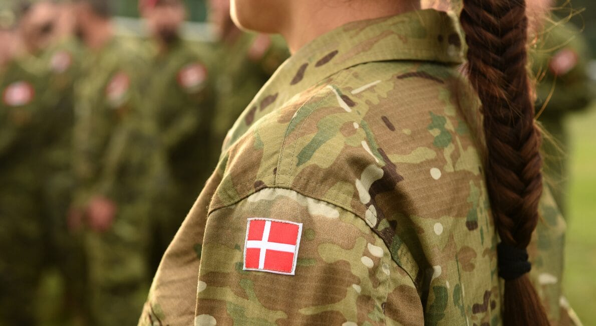 En person i militær camouflage med et dansk flagmærke på ærmet står i fokus med ansigtet vendt væk. Andre soldater, muligvis også kvinder, der opfylder værnepligt Danmark, er slørede i den udendørs baggrund.