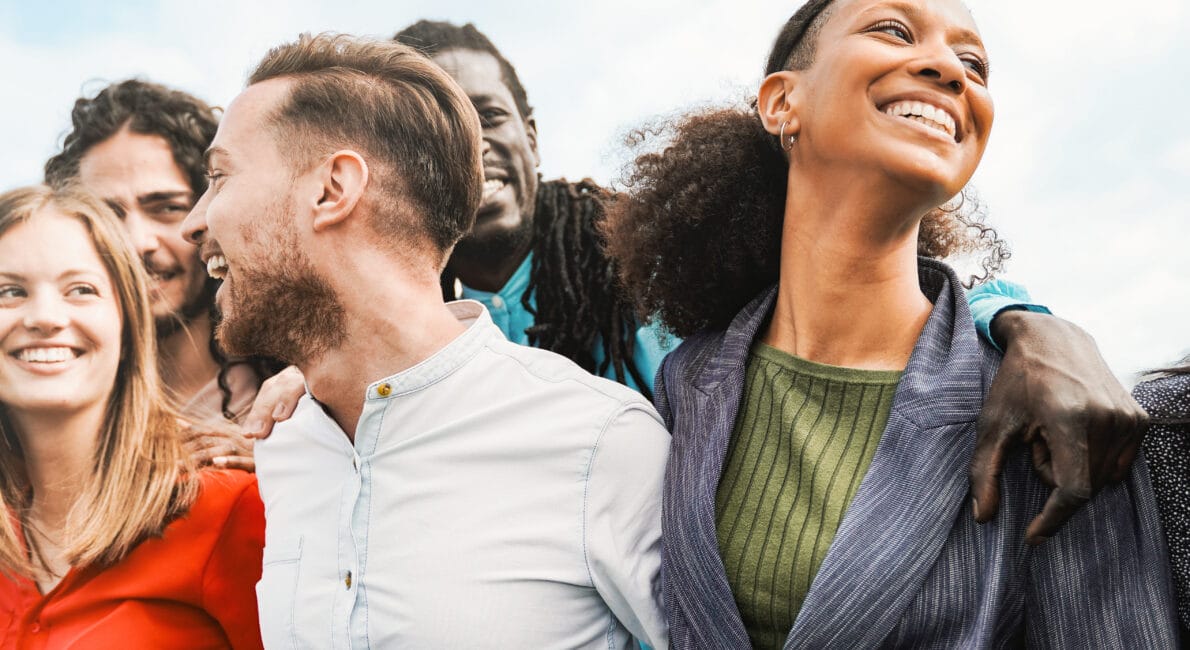 En mangfoldig gruppe unge voksne står tæt sammen udendørs, smilende og grinende, med blå himmel i baggrunden.