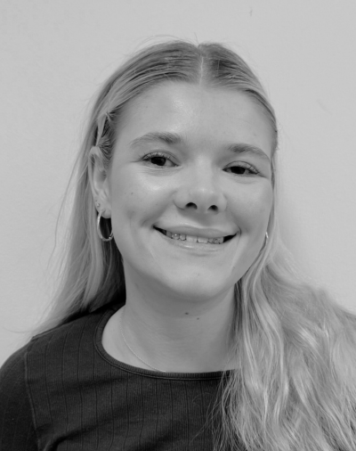 A young woman with long, light-colored hair smiles at the camera. She is wearing hoop earrings and a dark top. The photo is in black and white, and the background is plain.