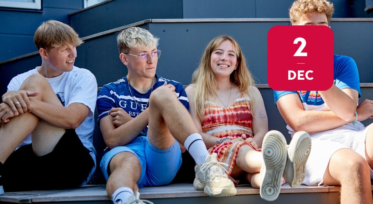 Fire teenagere sidder på en udendørs trappe og smiler og snakker. Den ene pige har et rødt batiktøj på, mens de tre drenge har afslappet sommertøj på. En rød firkant med 2 DEC er overlejret i øverste højre hjørne.