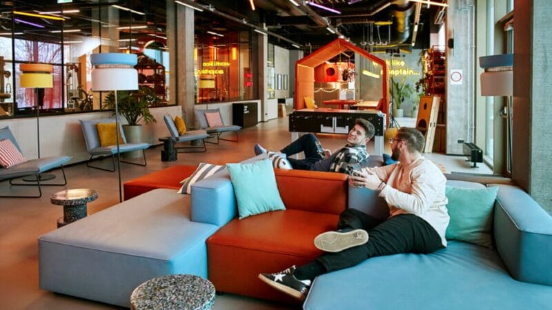 To personer sidder og taler, engageret i rådgivning, i farverige, modulopbyggede sofaer i en moderne, åben kontorlounge med levende belysning, legende indretning, et poolbord og store vinduer i baggrunden.