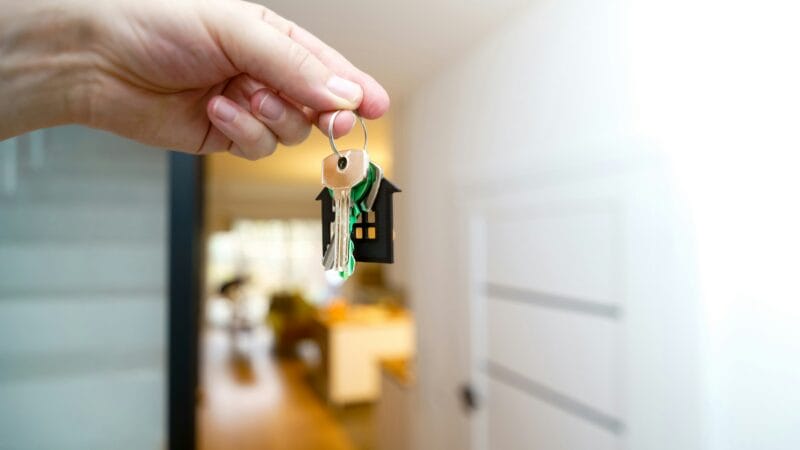 En hånd holder et sæt nøgler med en lille husformet nøglering i et stærkt oplyst, moderne boliginteriør, hvilket antyder, at man ejer et hjem eller flytter ind.