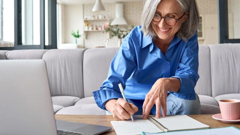 En smilende ældre kvinde med gråt hår og briller sidder i en sofa og skriver i en notesbog på et sofabord ved siden af en bærbar computer og en kop te i en hyggelig stue.