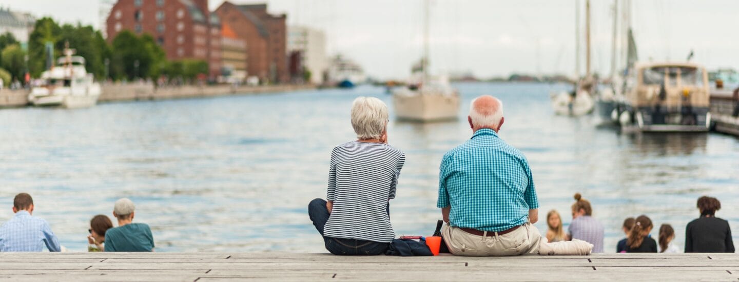 To ældre voksne sidder side om side på en træbrygge med udsigt til en havn med både og bygninger, omgivet af andre mennesker, der slapper af ved vandet på en klar dag.