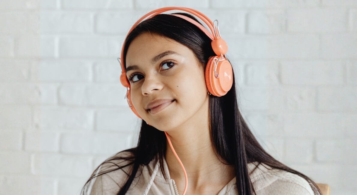 En ung kvinde med langt mørkt hår iført orange hovedtelefoner og en lys hættetrøje smiler blidt, mens hun kigger lidt til siden og står foran en hvid murstensvæg.