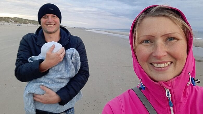 En kvinde i en pink jakke smiler til en selfie på en sandstrand, mens en mand i en sort hue og jakke står bag hende med en baby svøbt i et tæppe. Himlen er overskyet, og stranden er stort set tom.