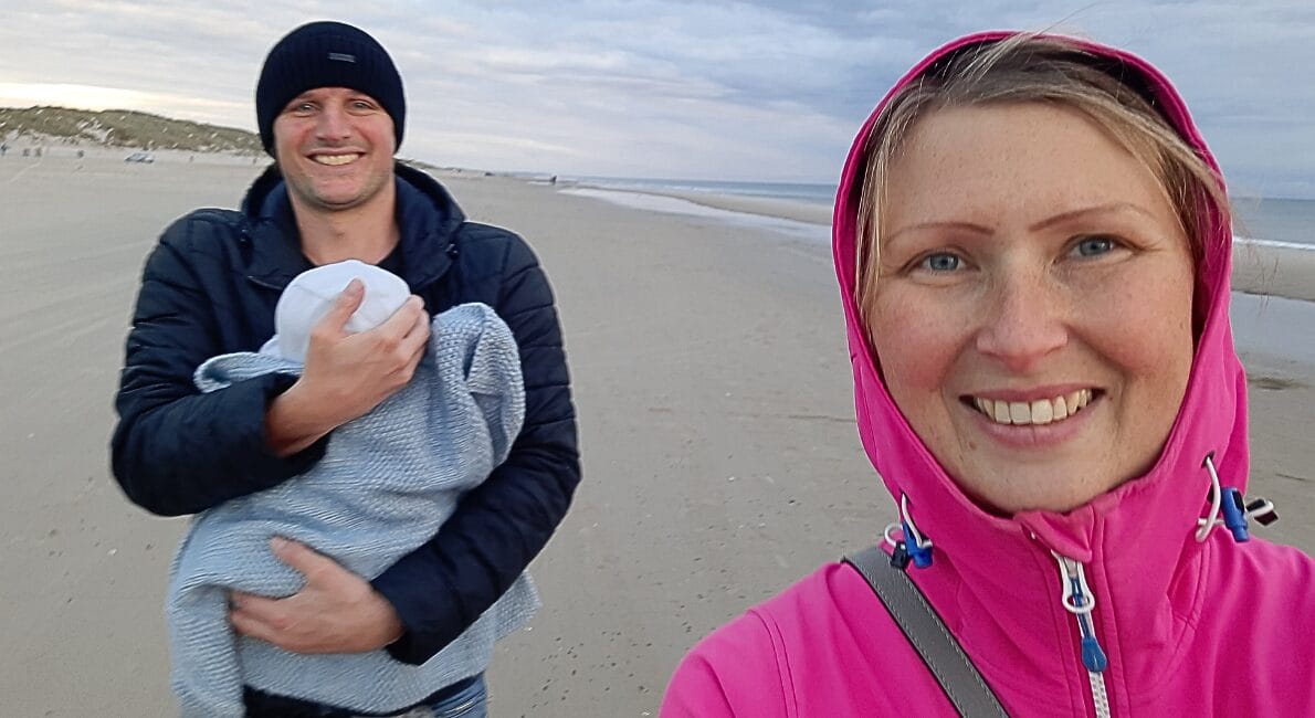 En kvinde i en pink jakke smiler til en selfie på en sandstrand, mens en mand i en sort hue og jakke står bag hende med en baby svøbt i et tæppe. Himlen er overskyet, og stranden er stort set tom.