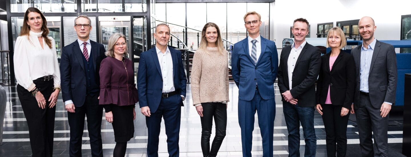 A group of nine experts stand in a modern office lobby with glass walls. Dressed in professional attire, they smile confidently at the camera. The floor is black and white tiled, and a staircase graces the background.