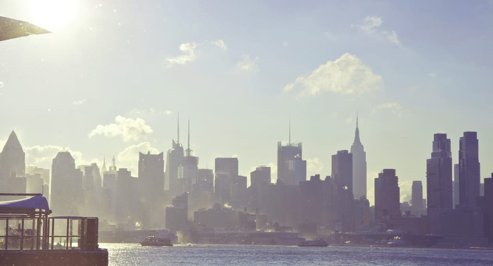 Billedet viser en byhorisont med høje bygninger i silhuet mod en lys, diset himmel. En stor vandmasse optager forgrunden, hvor et par både glider blidt. Sollys kaster et blødt skær over scenen og fremkalder drømme om at tage på job i udlandet for nye muligheder i udlandet.