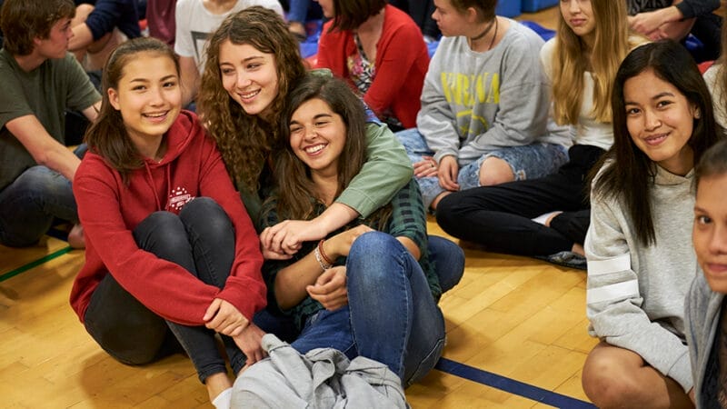En gruppe smilende teenagere sad på et gymnastiksals gulv. Tre piger i centret nusser og griner. Andre teenagere sidder omkring dem, afslappet klædt. Atmosfæren er venlig og afslappet.