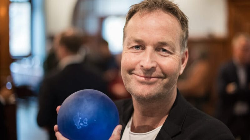 En person, der smiler og holder en blå kugleformet genstand, muligvis en bowlingkugle, indendørs. Baggrunden er sløret og viser andre mennesker og træpaneler.
