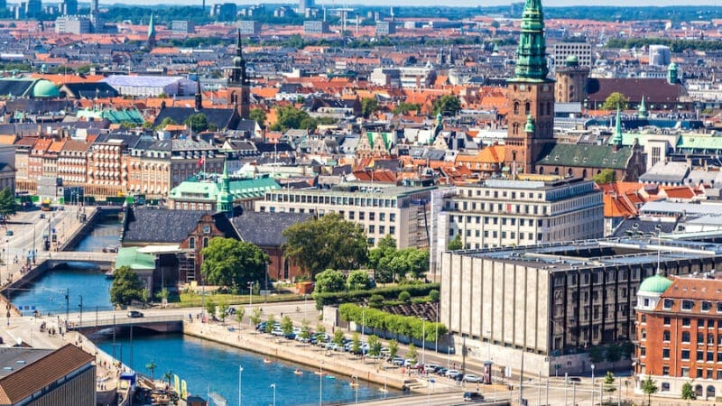 Luftfoto af et bybillede med historiske bygninger, kanaler og grønne spir, der minder om dem, du måske forestiller dig, når du flytter tilbage til Danmark. Skyline kan prale af strukturer med røde tag sammen med nogle få moderne bygninger under den klare blå himmel, hvilket skaber en levende og travl scene.