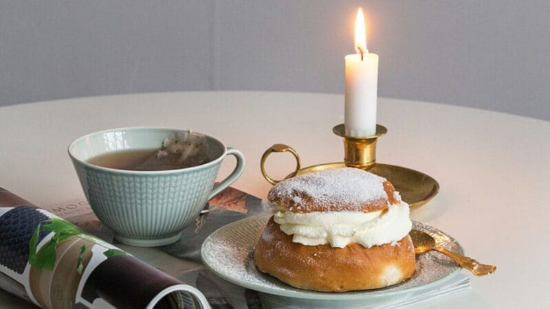 En hyggelig scene med en kop te ved siden af et flødefyldt wienerbrød tilstrøet med pulveriseret sukker, hvilende på en lille tallerken med en ske. Et tændt lys i en messingholder og et sammenrullet magasin er placeret i nærheden på et rundt bord.