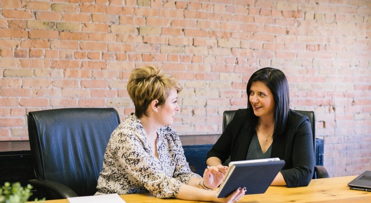 Two women are seated at a wooden conference table in an office with a brick wall. One woman is holding a tablet and discussing it with the other. Both appear engaged and smiling. Two black chairs and a plant are visible.