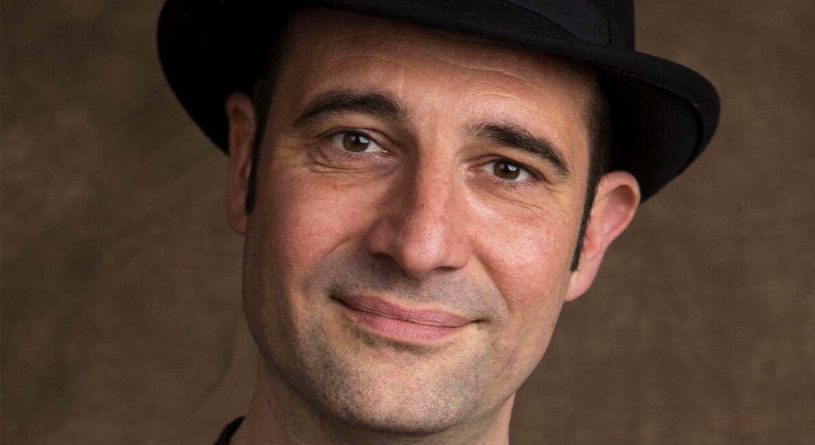 En person, der bærer en sort hat og et lille smil, poserer mod en brun baggrund.