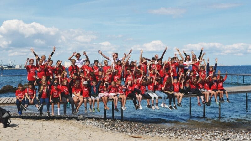 En stor gruppe mennesker iført røde skjorter, måske fra et glædeligt teambuildingjob-retræte, sidder på en træmole og løfter muntert armene. De er ved stranden med havet og en lysende blå himmel som baggrund.