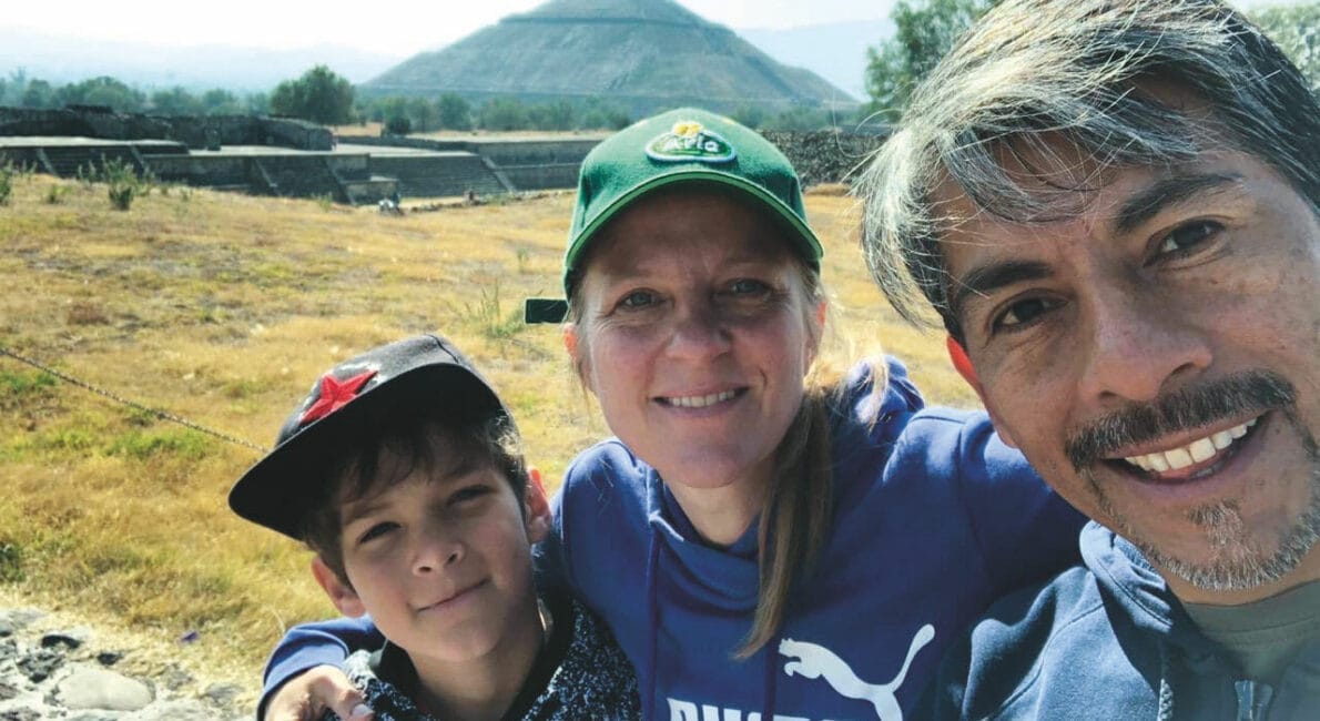 En familie på tre, der smiler til en selfie udendørs, med en gammel pyramide i baggrunden. Barnet har en stjernehat på, kvinden en grøn kasket, og manden en grå jakke. Landskabet er græsklædt med en stensti.