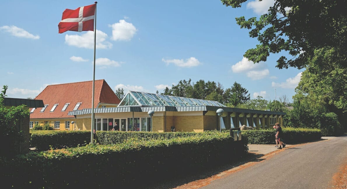 En murstensbygning med glastilbygning er omgivet af grønt og træer under en blå himmel. Et dansk flag står på en flagstang foran, og en person går langs en nærliggende vej.