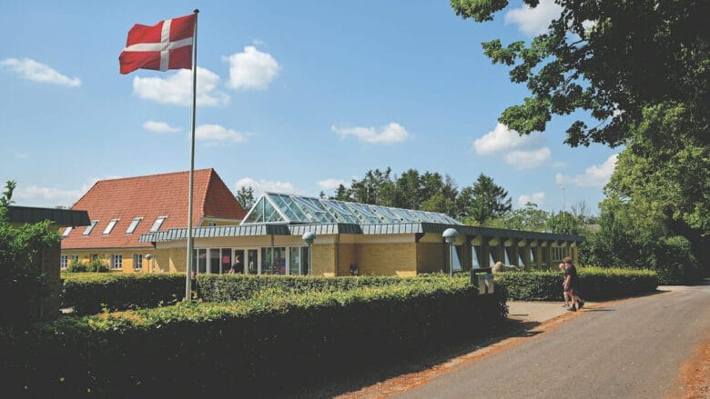 En murstensbygning med glastilbygning er omgivet af grønt og træer under en blå himmel. Et dansk flag står på en flagstang foran, og en person går langs en nærliggende vej.