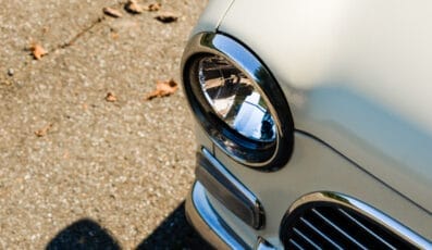 Close-up of the front corner of a vintage car, focusing on a round chrome headlight and part of the grille. The car glistens under the sun on an asphalt road sprinkled with brown leaves, capturing timeless style—much like discovering rare discounts in an unexpected place.