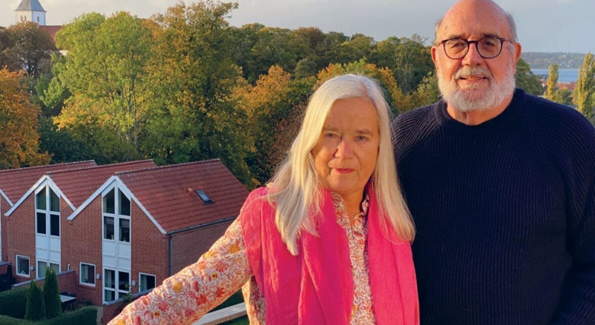 Et ældre par står på en altan med en naturskøn udsigt. Kvinden har langt hvidt hår, bærer en blomstret skjorte og et lyserødt tørklæde. Manden har skæg, briller og er iført en mørk sweater. Bag dem er træer, huse og en overskyet himmel.