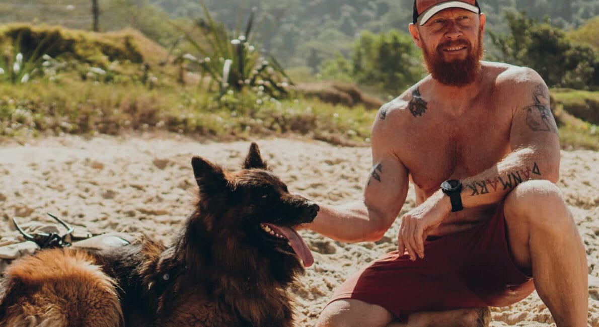 En mand uden skjorte iført kasket og røde shorts smiler, mens han knæler ved siden af en schæferhund på en sandstrand. Hunden sidder ved siden af en blå skål. Frodige, grønne bakker er synlige i baggrunden.