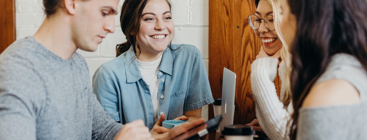 Fire personer sidder ved et bord med notesbøger og en bærbar computer og deltager i en livlig diskussion. Når hvert medlem bidrager med ideer, virker de afslappede og smilende.