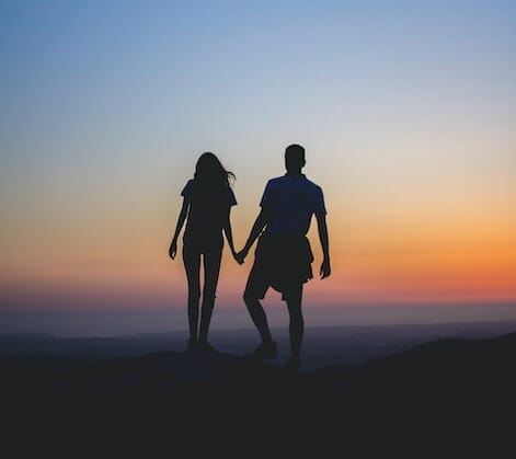 Silhuetbillede af to mennesker, der holder hinanden i hånden mod en farverig solnedgangshimmel, som om de var frosset i et øjebliks sindsro. Horisonten strækker sig ud over dem og kaster et roligt skær svarende til færdiggørelsen af en følelsesmæssig sagsbehandling, hvilket skaber en unægtelig fredelig og romantisk atmosfære.