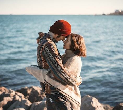 Et par står på klipper ved havet og omfavner hinanden tæt. Indhyllet i hyggelige trøjer og en rød hue deler de et intimt øjeblik med rørende pander, på baggrund af blåt vand og en klar himmel – naturens egen blide sagsbehandling for sjælen.