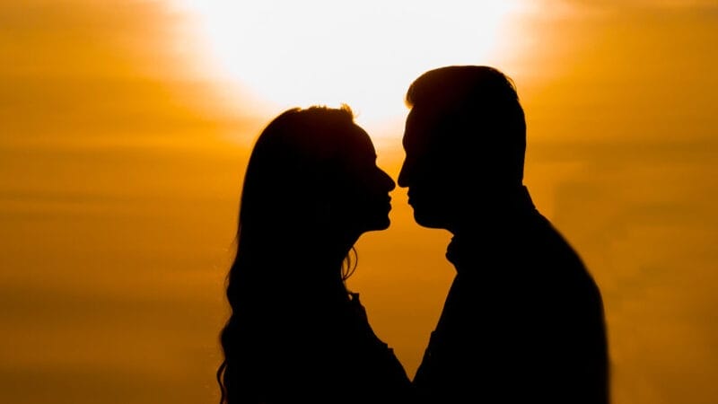 Silhuet af et par, der står over for hinanden mod en levende solnedgang, der inkarnerer familiesammenføring. Solen skaber et varmt skær omkring dem og fremhæver deres profiler, når himlen skifter gennem en gradient af orange og gule nuancer.