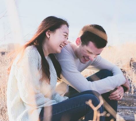 En kvinde og en mand sidder udendørs i tørt græs, begge iklædt trøjer og jeans. Med smil lyse som den klare himmel ovenover læner de sig frem, deler et glædeligt øjeblik, deres latter er så blid som brisen - en ubesværet scene, der ikke kræver sagsbehandling.