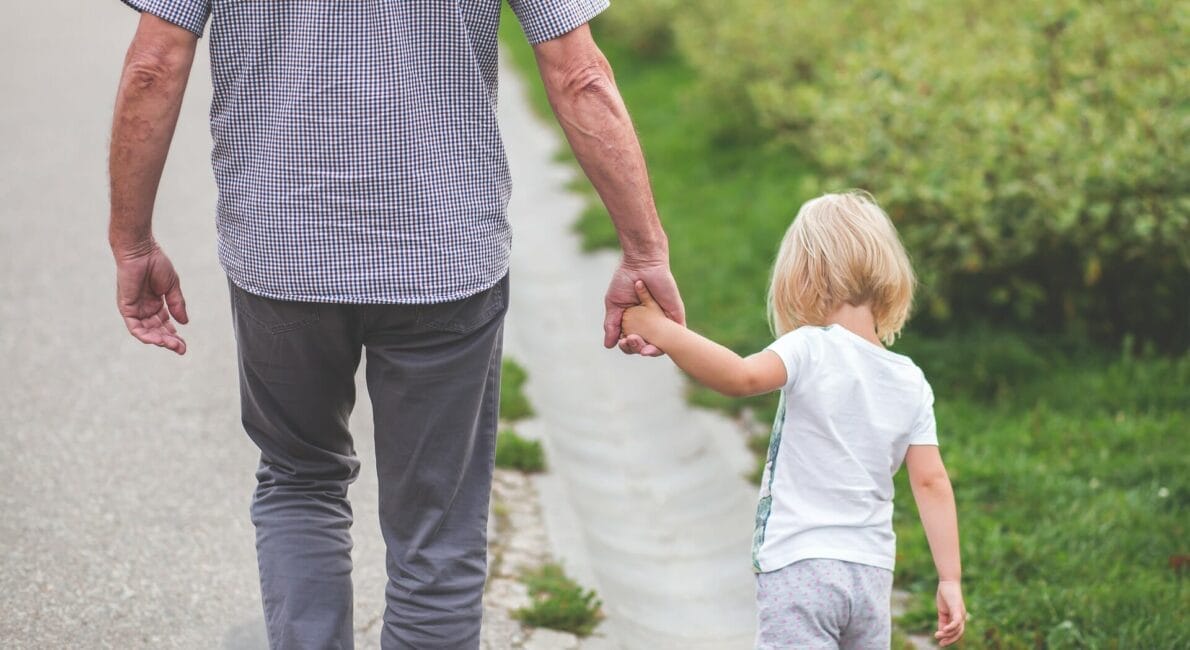En mand og et lille barn går hånd i hånd ad en sti. Barnet har lyst hår og er iført hvid skjorte og shorts. Manden er iført en ternet skjorte og grå bukser. De er omgivet af grønne områder.
