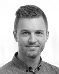 Portræt af en person med kort hår og lyst skæg, iført en skjorte med knapper. De ser direkte på kameraet med et neutralt udtryk. Baggrunden er almindelig og lysfarvet, hvilket skaber et professionelt udseende.