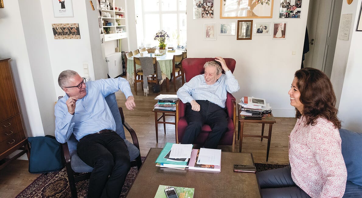 Tre personer sidder og smiler i en hyggelig stue og går i snak. To mænd i lyseblå skjorter sidder i lænestole, mens en kvinde i en lyserød top sidder på en sofa. Rummet har en bogreol, billeder og et spisebord i baggrunden.