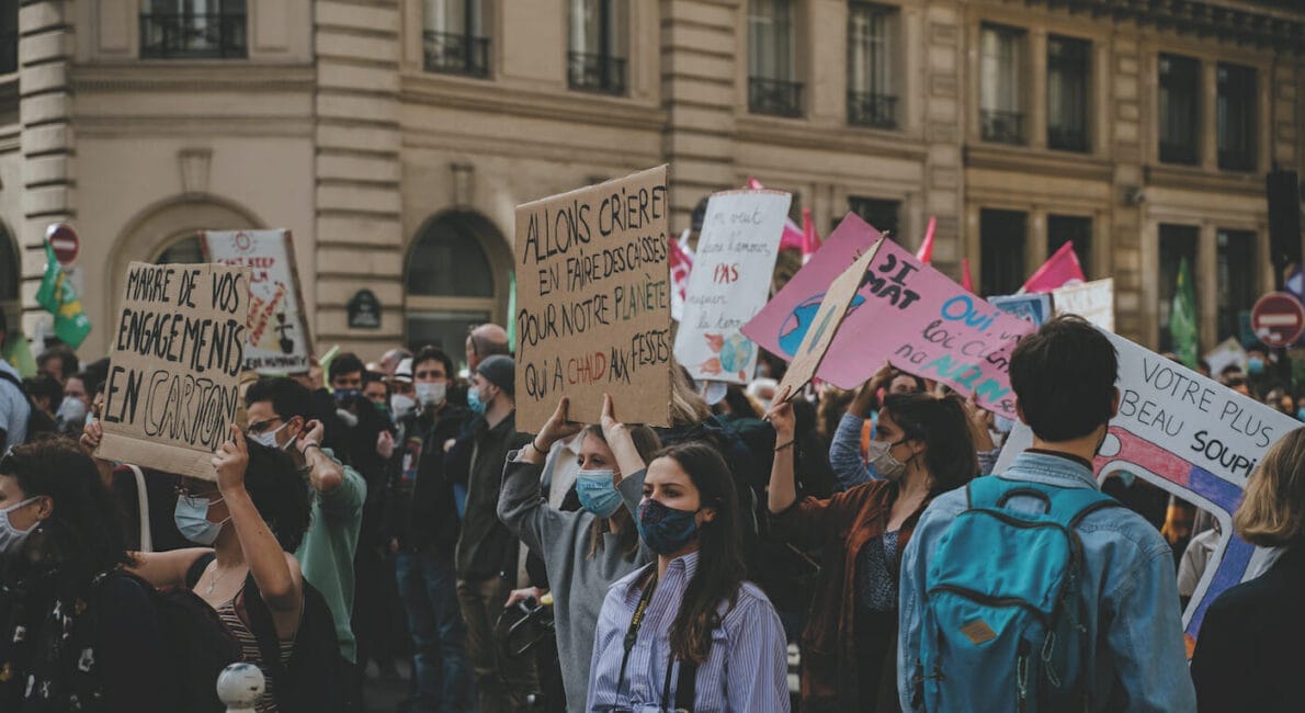 En gruppe mennesker iført masker er samlet i en gadeprotest. De holder forskellige skilte op med fransk tekst. Baggrunden viser en bygning med klassisk arkitektur.