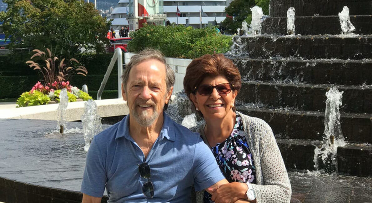 Et smilende par sidder foran et stort springvand. Manden er iført blå skjorte og briller om halsen, mens kvinden er i mønstret top og sweater, der begge fremstår glad og afslappet på en solskinsdag.