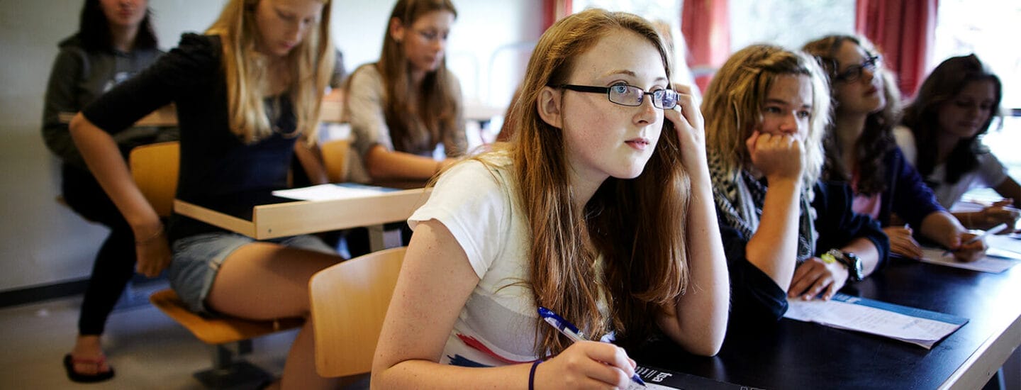 En gruppe studerende i et klasseværelse, der sidder ved skriveborde med notesbøger og kuglepenne. En pige med briller og rødt hår glæder sig opmærksomt, mens andre er fokuserede på deres noter. Rummet har lyst naturligt lys fra vinduerne.