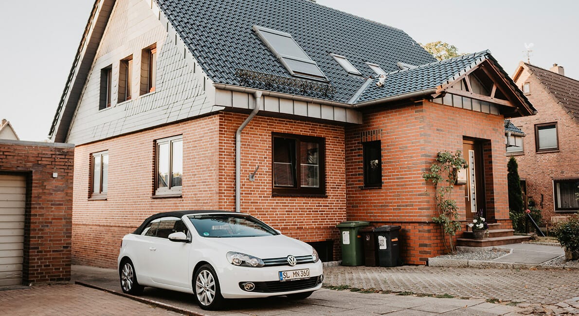 En hvid cabriolet parkeret foran et to-etagers murstenshus med kvistvinduer og et blåt tegltag. Indkørslen har et par skraldespande, og garagen er delvist synlig til venstre.