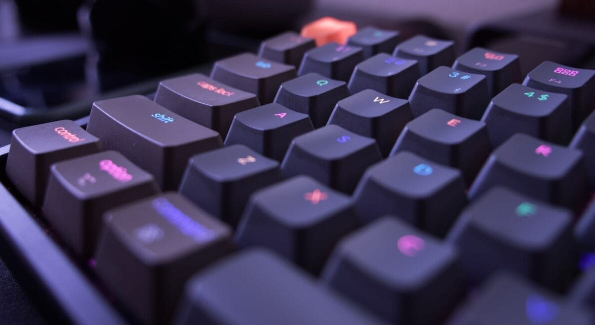 Nærbillede af et mekanisk tastatur med sorte taster. Tasterne har RGB-belysning, der viser forskellige farver. "ESC"-tasten er unikt orange. Fokus er på de vinklede taster, hvilket skaber en dynamisk visuel effekt.
