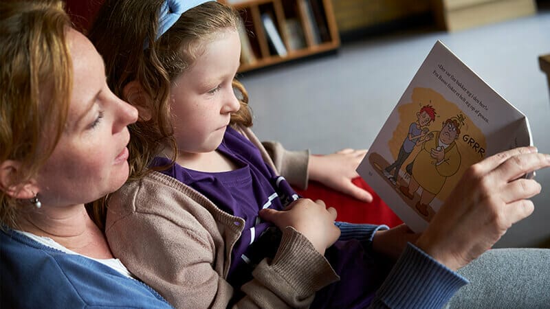 A woman and a young girl are sitting together on a couch, reading a childrens book. The girl is holding a stuffed toy, and they appear focused on the book. The room has soft lighting, with bookshelves visible in the background.